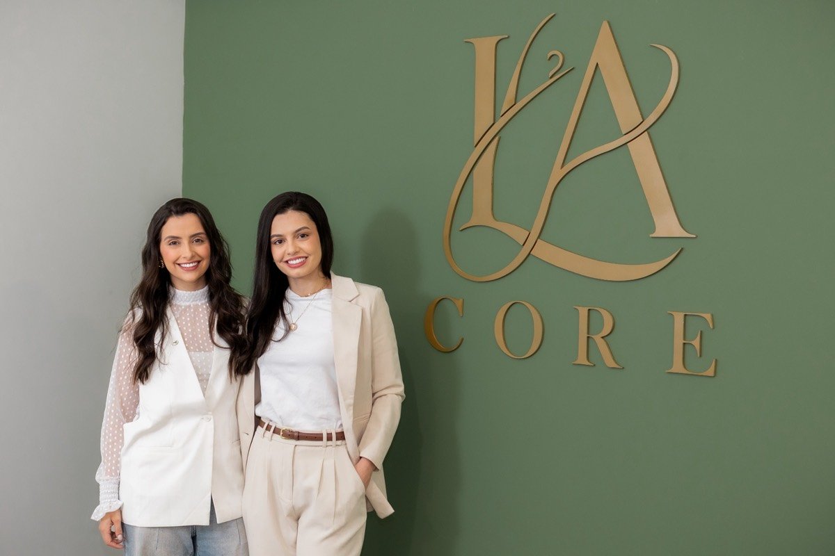 As fisioterapeutas Lorena Araújo e Anny Caroline ao lado do logo da LA Core na parede verde